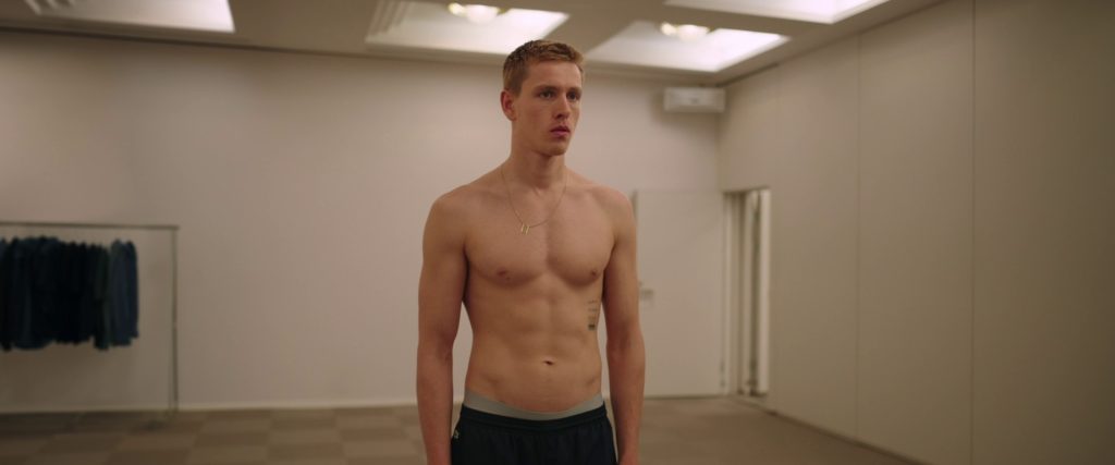Harris Dickinson torse nu dans une pièce blanche au début de Sans Filtre de Ruben Östlund.