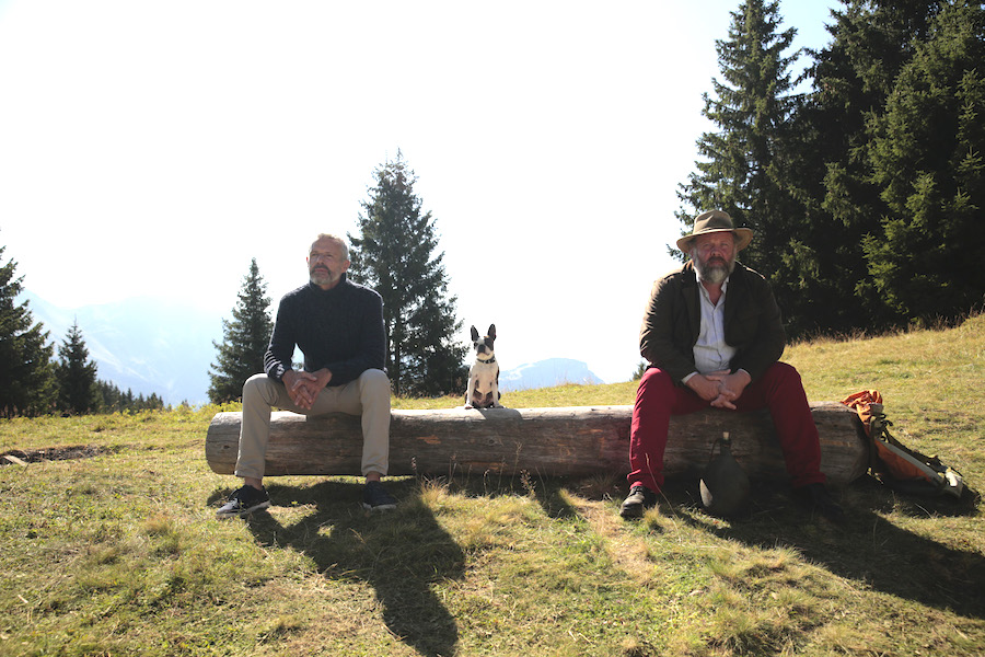 Lambert Wilson et Grégory Gadebois sur un banc à la montagne avec un chien au milieu.