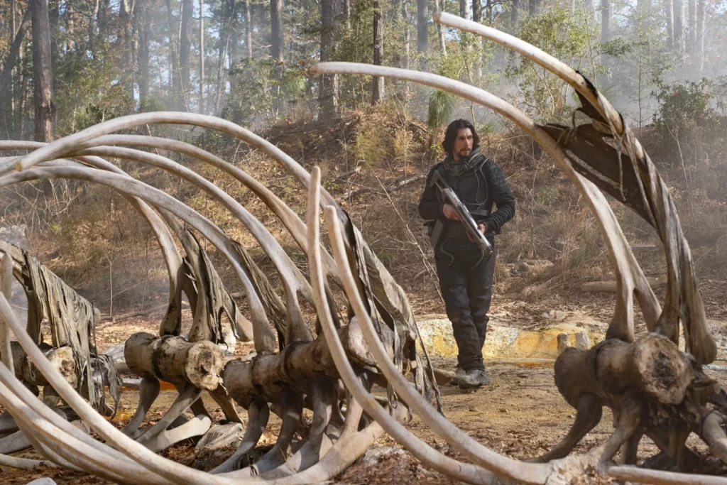 Adam Driver et son arme au milieu des ossements de dinosaures dans la forêt.
