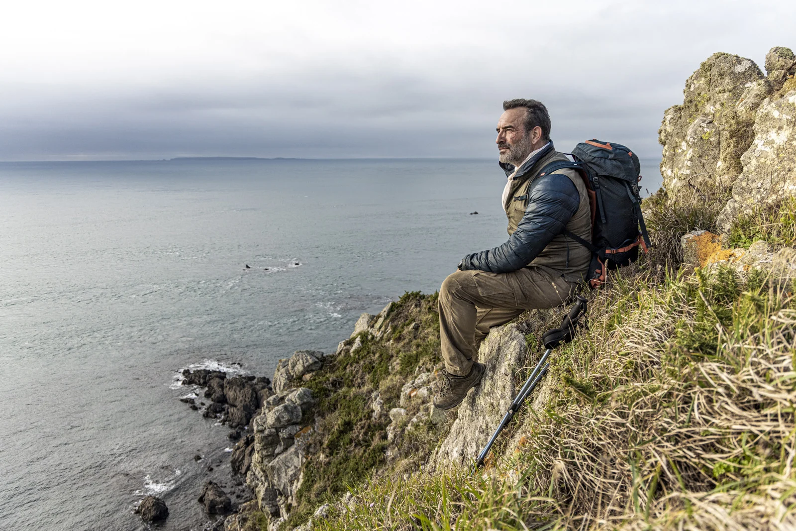 Jean Dujardin assis sur un rocher face à la mer dans Sur les chemins noirs.