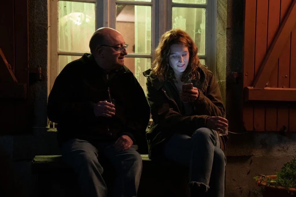 Michel Blanc et Julia Piaton regardent le téléphone en buvant une bière devant la maison la nuit.