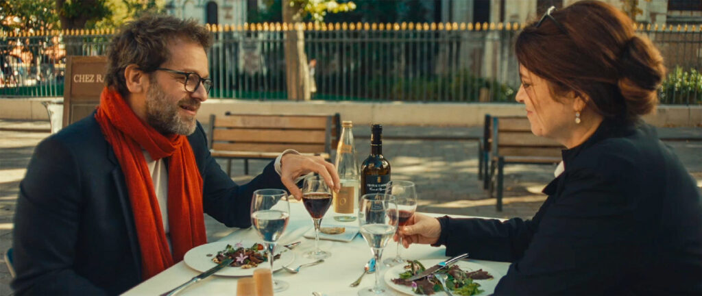 Jonathan Zaccaï et Agnès Jaoui à table en terrasse écharpe rouge et verres de vin.