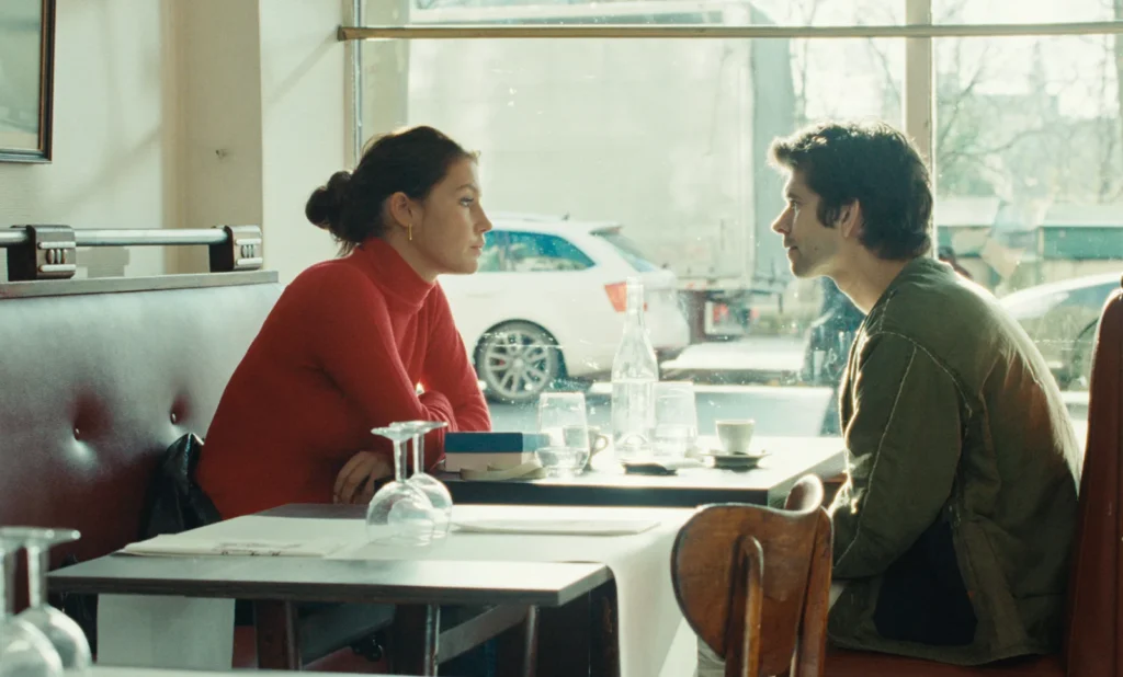 Adèle Exarchopoulos pull rouge face à Ben Whishaw au café de profil.