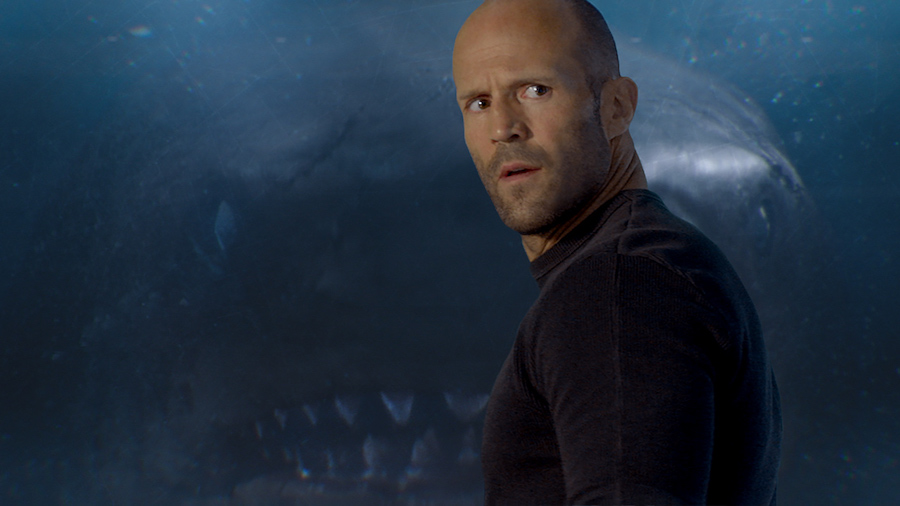 Jason Statham de trois quarts avec u un requin dans le dos derrière la vitre.