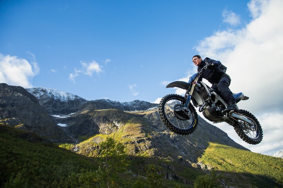 Tom Cruise saut à moto en montagne tenue noire devant le ciel.